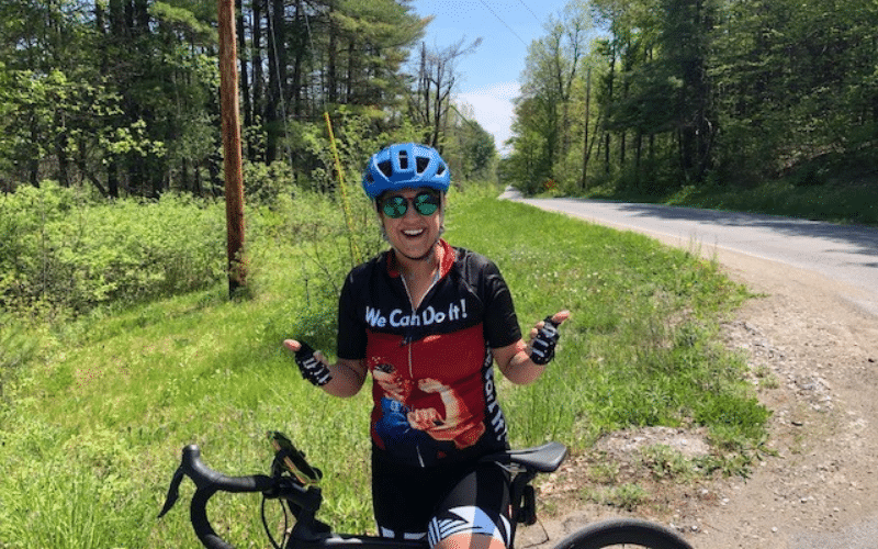 Jenna, A white woman, is standing behind a bike, wearing a "We Can Do It!" biking shirt. She stands in front of a road and deciduous tree forest.