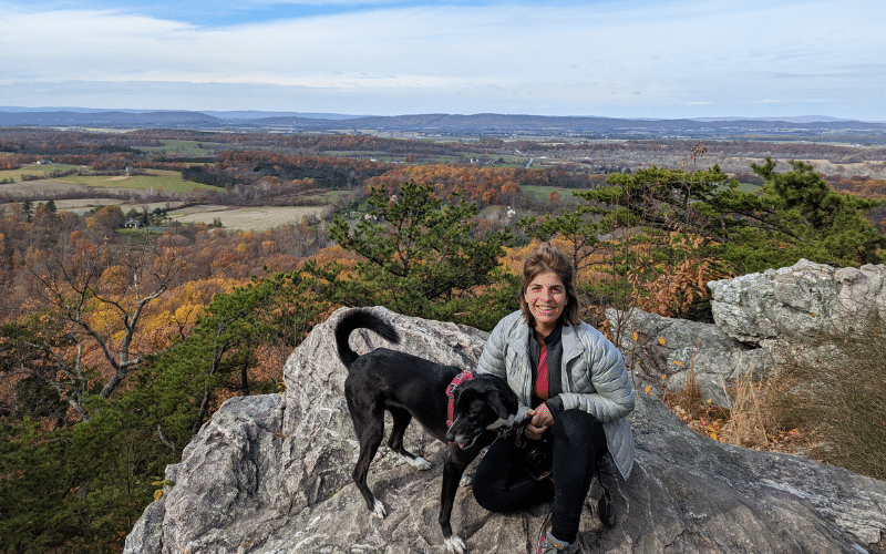 Karina, a woman with brown hair, crouches on a rock ledge with a black dog with white paws. The rock overlooks a beautiful background of trees, mountains, and sky.