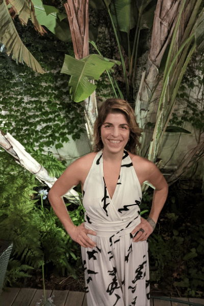 Karina stands in front of some plants. She is wearing a white jumpsuit and smiling.