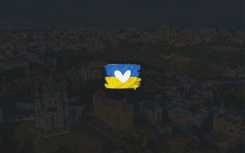 Image description: White heart over small ukrainian flag, over dark image of Kyiv cityscape