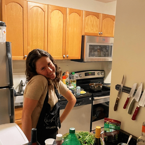 Rachel stands in a kitchen, wearing an apron, smiling and resting her head on her right shoulder.