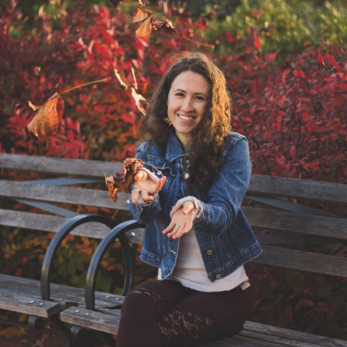 Rachel sits on a park bench surrounded by autumn leaves and is holding her hands out towards the camera