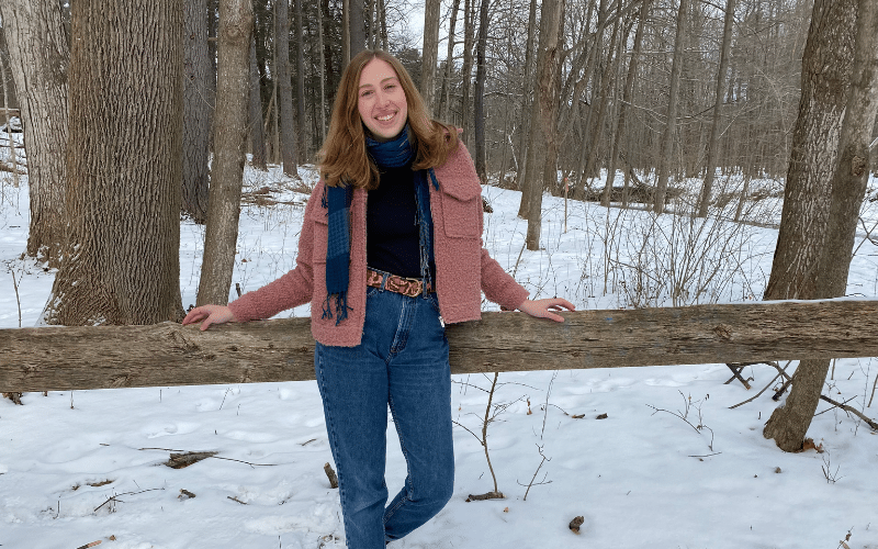 Hannah, a white woman, leans against a wooden fence. The ground is covered in snow and there are trees in the background