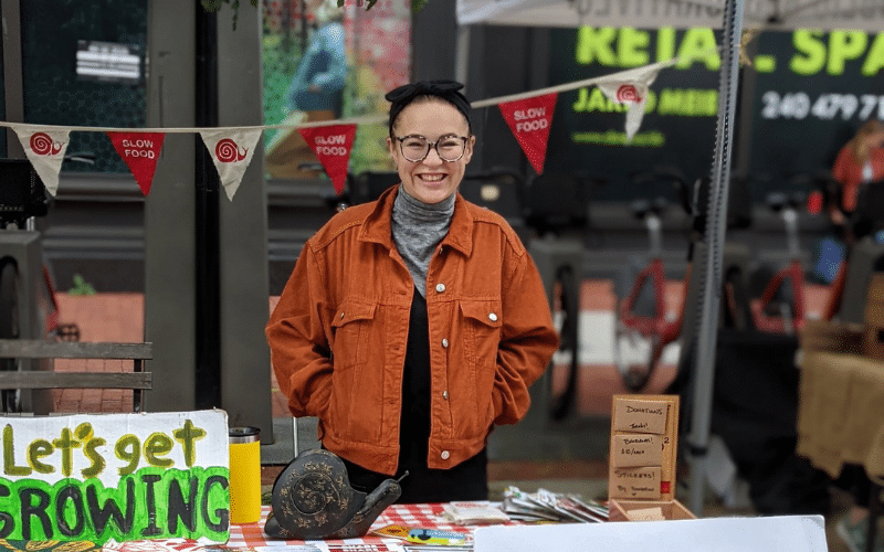 Hannah, a white woman, stands at a market behind a table that says "Let's get growing". She is smiling and wearing an orange jacket.