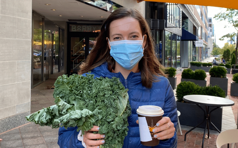 Abby smiles at the camera, wearing a blue mask and holding kale in one hand and a hot to-go drink cup in the other hand