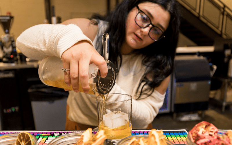 Rivka had dark hair and wears glasses. She is standing behind a bar pouring an old fashioned from a cocktail shaker into a glass with an ice cube
