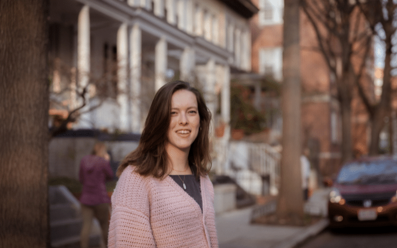 Abby wears a pink cardigan and smiles at the camera in front of a DC block of rowhouses in the background