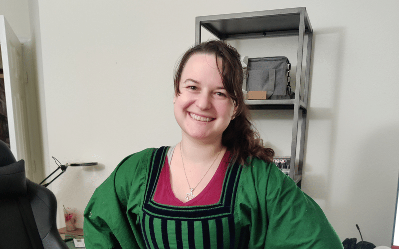 Emily, a white woman, smiles and stands in a green and black handmade period dress