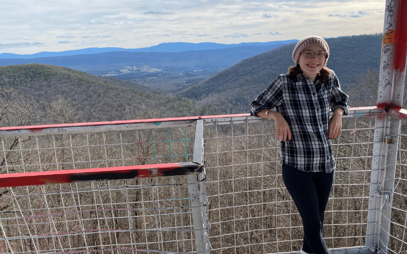 Sam, a light skinned woman, stands at a lookout point overlooking a picturesque mountain range