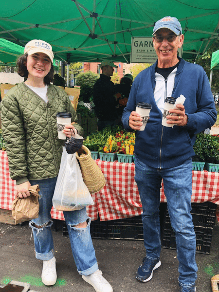 Becca and an older white man stand at a farmer's market holding shopping bags and coffee. 