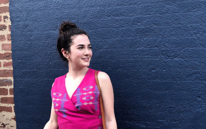 Becca, a white woman, stands in front of a black brick wall wearing a hot pink top and smiling towards something off camera to the side.