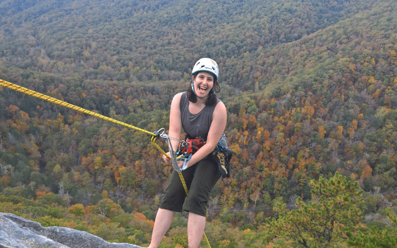 Wendy sticking her tongue out and repelling over the side of a cliff with a forest that is just starting to turn to fall colors below her.
