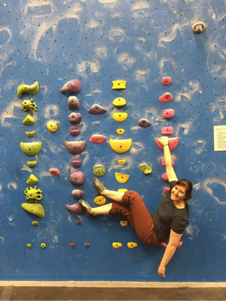 Wendy hangs almost horizontally off of an indoor climbing wall using only one are 
