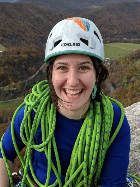 Wendy stands on top of of cliff wearing a white climbing helmet and has a large amount of bright green rope resting around her neck