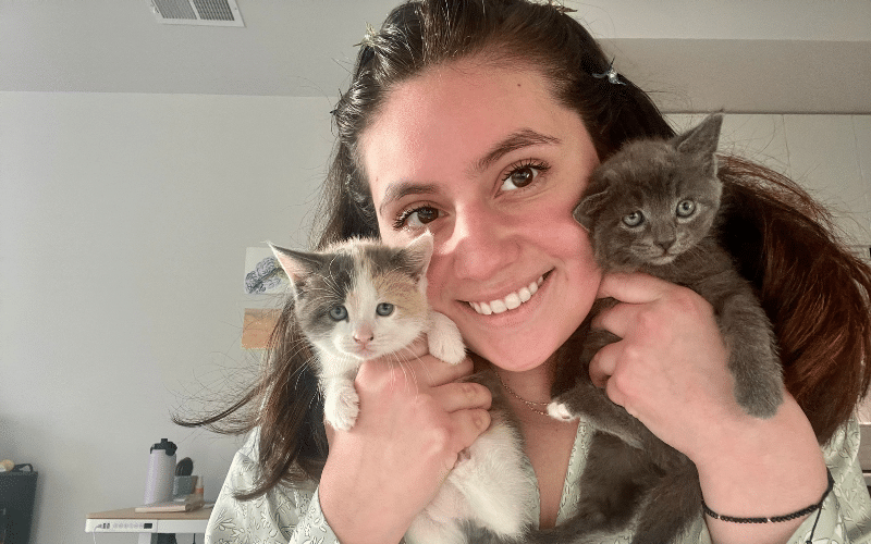 Yaniza holds two adorable tiny kittens, one gray and one mostly white, and smiles.