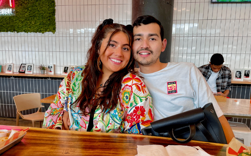 Yaniza, wearing a brightly colored pattern top, and her husband sit together at a restaurant. 