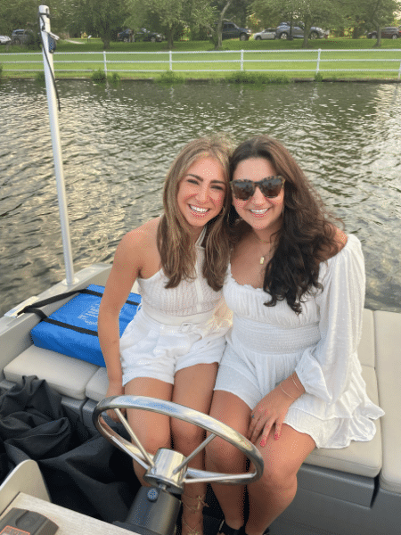 Sofie and a brunette woman sit in a boat behind the steering wheel in all white outfits.