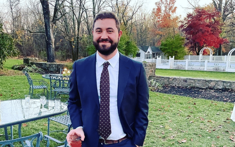 Jonathan, a white man with dark brown short hair and a close beard, wears a navy blue suit and stands in front of a well manicured garden and lawn.