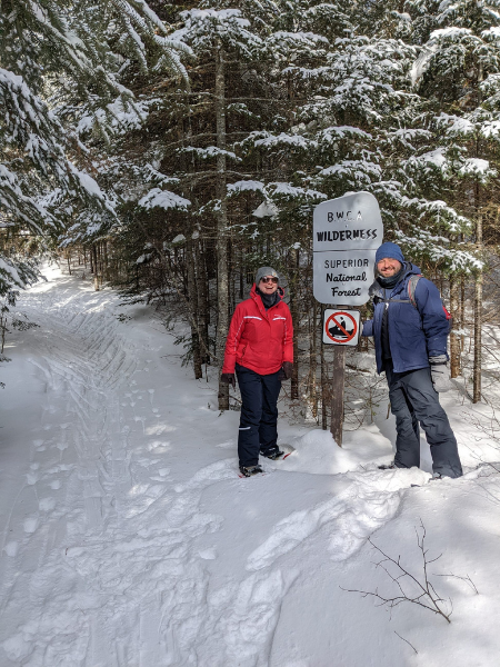 Jonathan and another person stand in the woods in heavy snow in front of a sign for a trail.