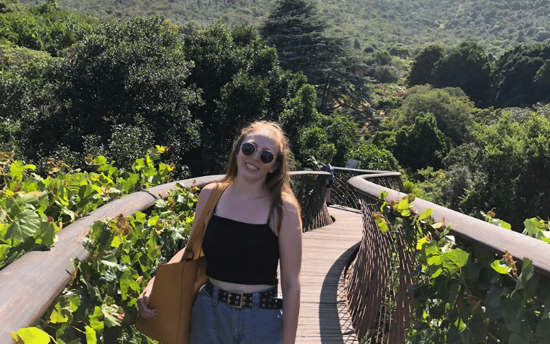 Leah, a white woman, smiles in a black top, jean shorts, and sunglasses. She stands on a winding wooden bridge in the middle of some luscious green mountains.