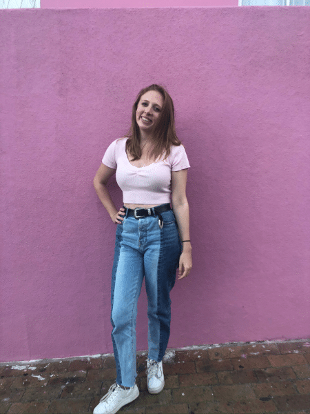 Leah poses in front of a pink wall. She is wearing multicolored jeans and holds one hand at her waist.