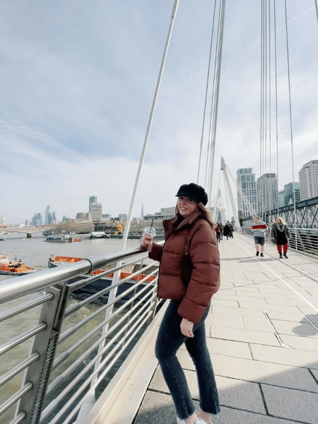Leah stands on a modern bridge holding a cup of coffee and wearing a very puffy coat.