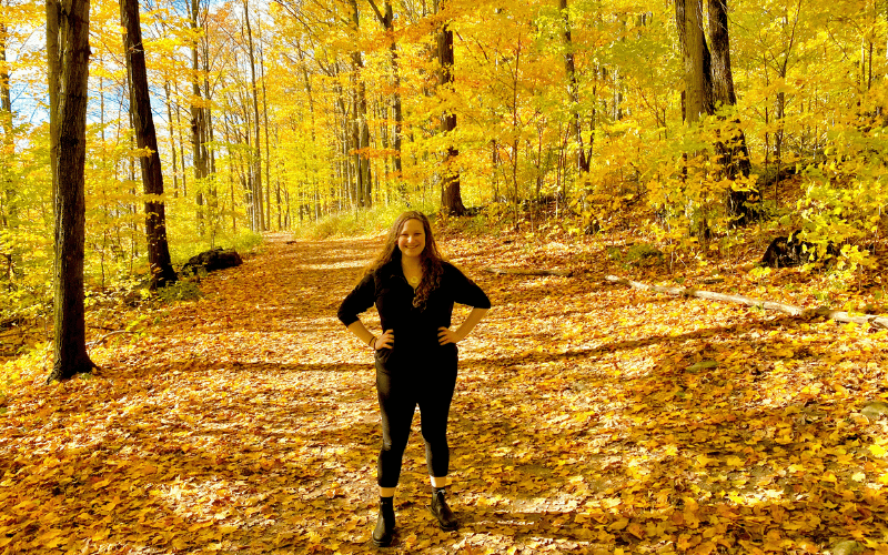 Rachel stands in the middle of a forest thats leaves have turned bright yellow.