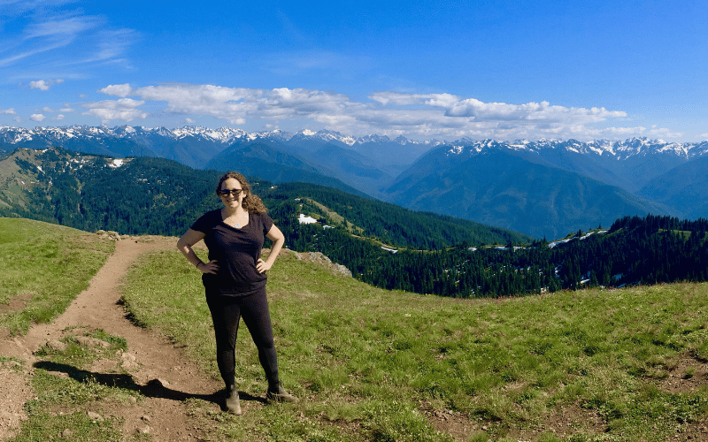 Rachel stands on a green spot on top of a mountain, overlooking a large snowcapped maintain range in the distance