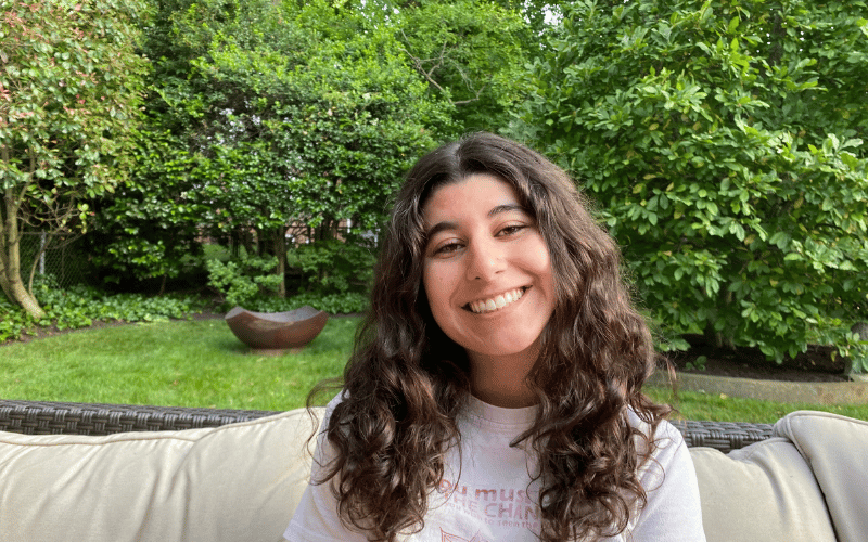 Tali, a white woman with curly brown hairs, sits on a couch in a green backyard, smiling. 