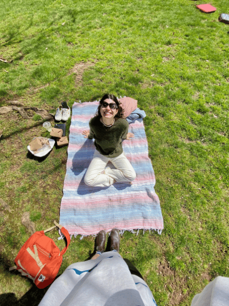 Talia sits on the ground on a picnic blanket looking upwards at the camera with a big smile