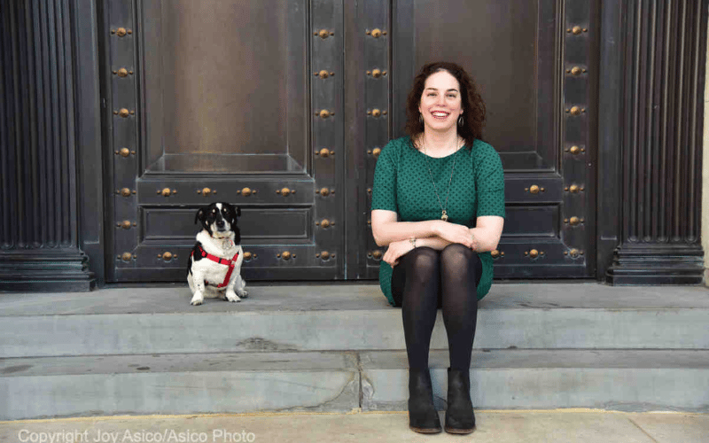 Glenna, a white woman with curly brown hair, sits on a small set of steps next to her very cute black and white dog.