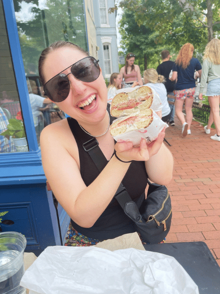 Allyson sits at a table outside, holding up her bagel sandwich to show the beautiful filling.