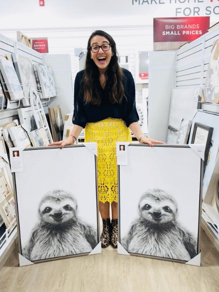 Alana smiles widely as she holds two identical sloth photographs in the aisle of a store.