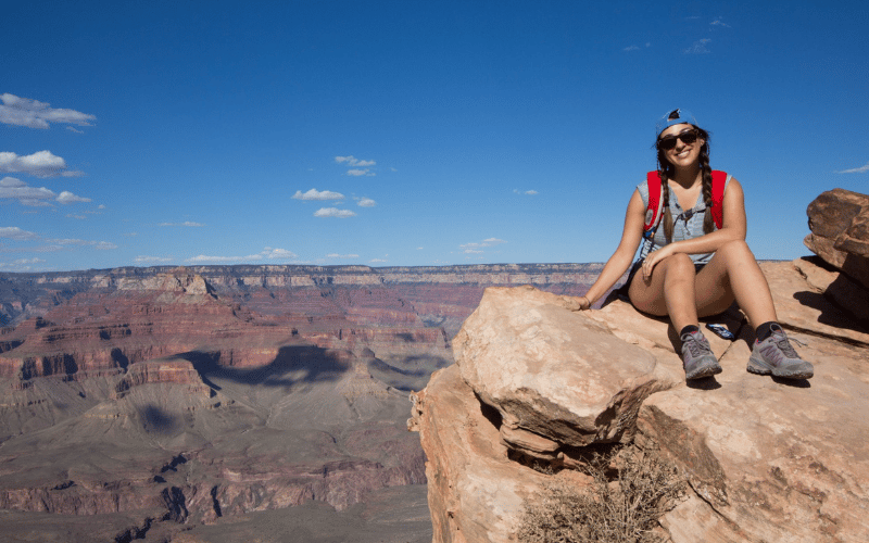 Alana sits in hiking gear on the edge of a rock overlooking a massive canyon.