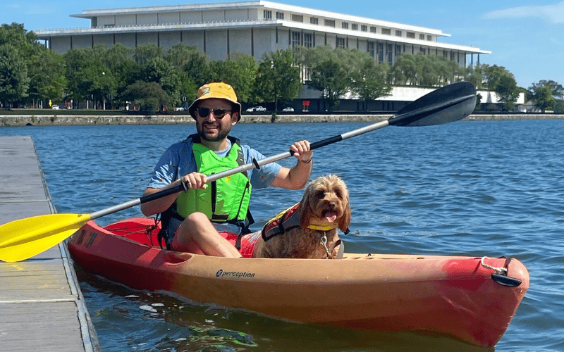 A white man, Michael, and his brown fluffy dog sit in a Kayak on the Potomac river with a large concrete building in the background