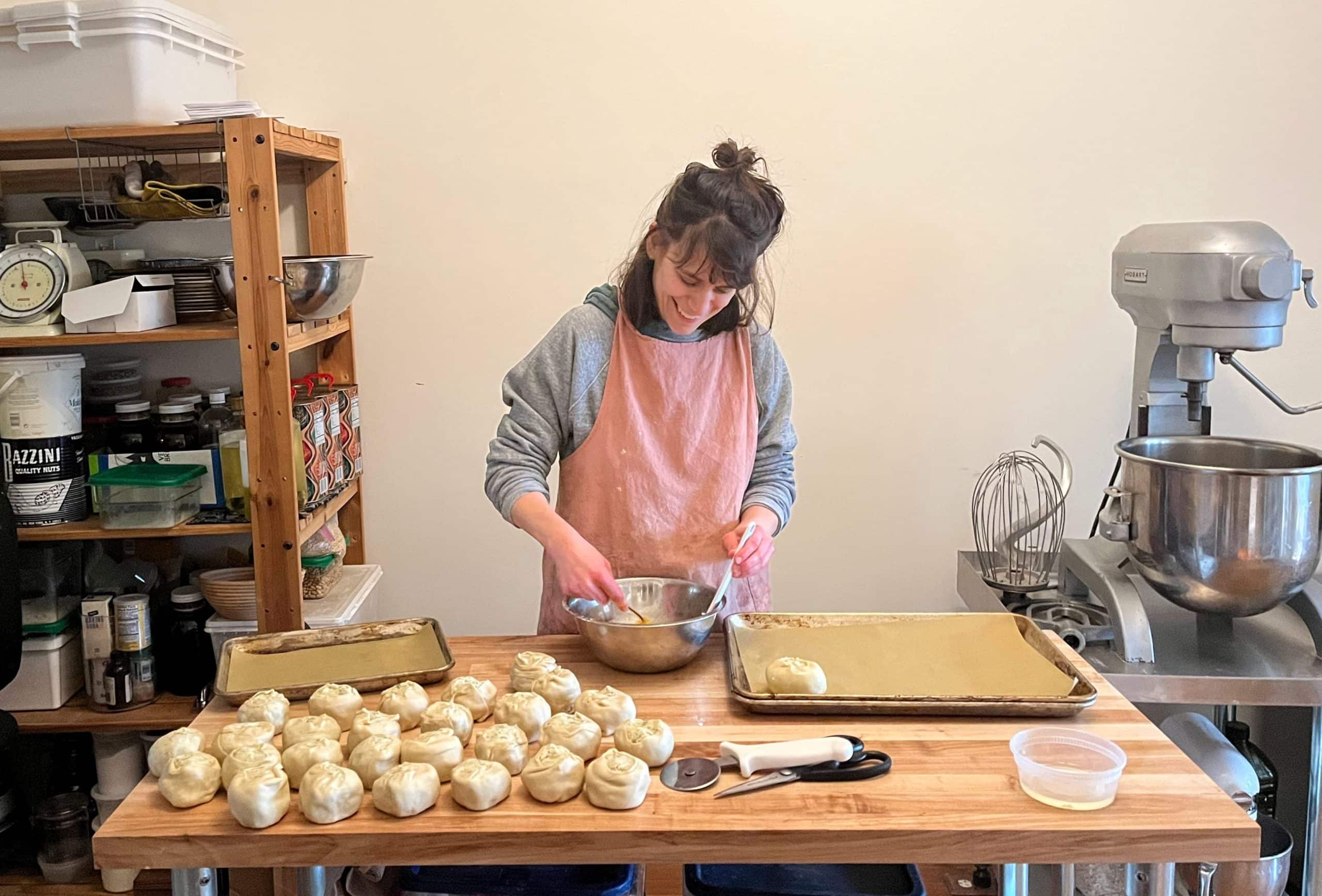 Image: woman in apron standing behind baker's table mixing dough with a dozen unbaked rolls in front of her