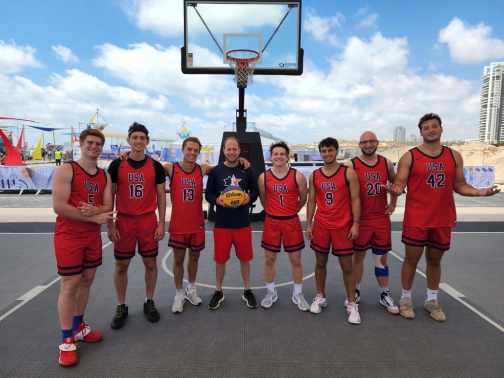 Michael Shapiro poses with the USA 3x3 basketball team under a basketball hoop on an outdoor court. 