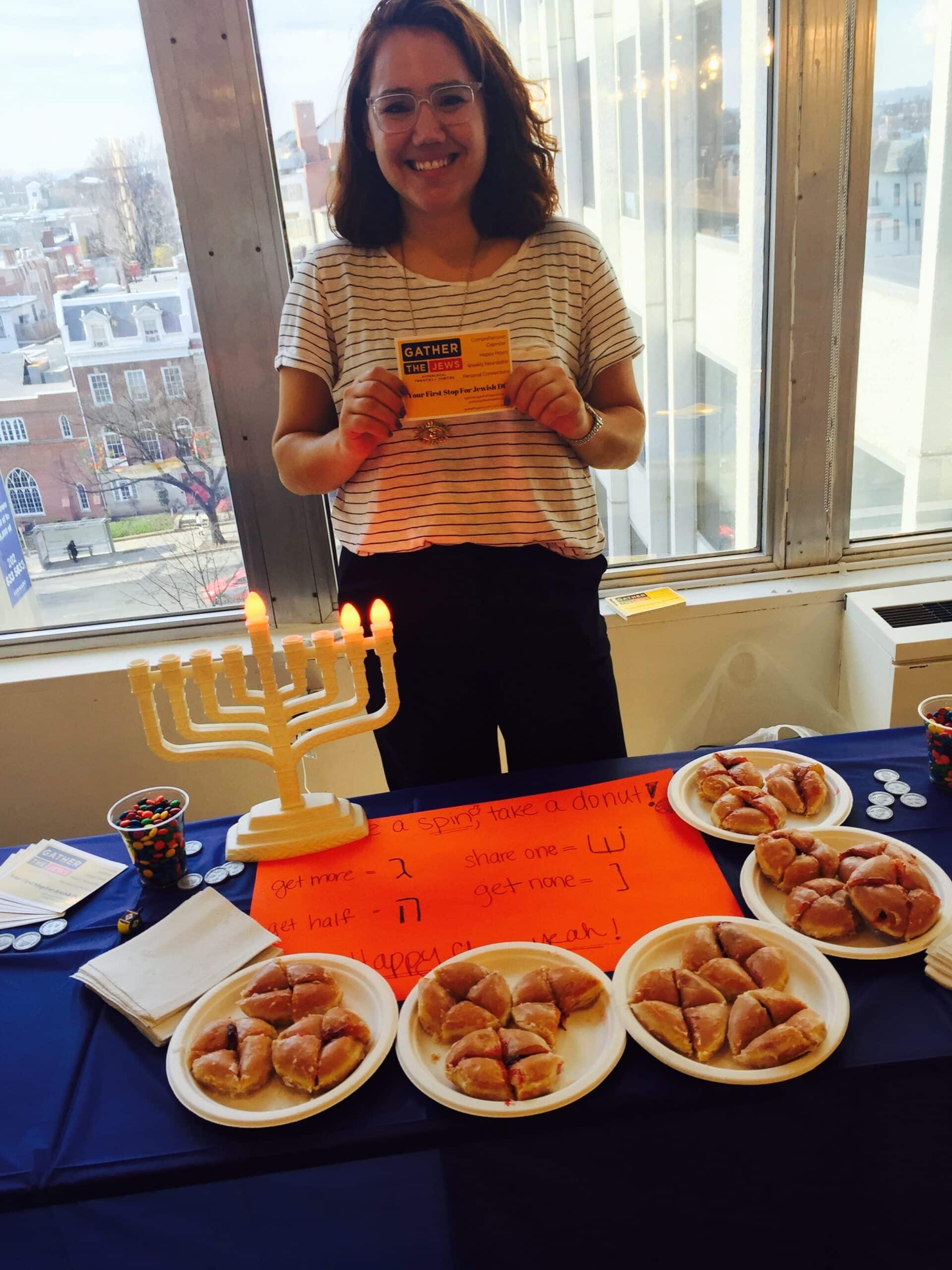 Jackie stands behind a table at a Gather event, fronted by a menorah, an orange sign, and plates of food. 