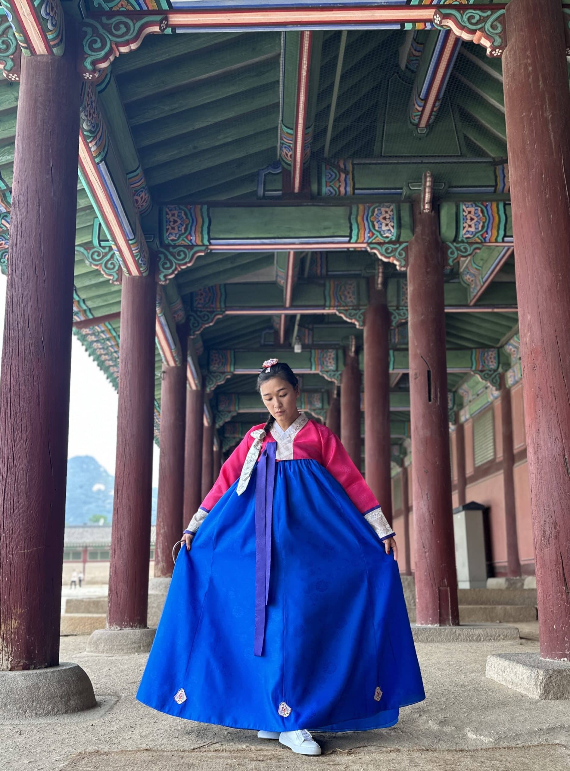Liz is standing in the outdoor hallway of Gyeongbokgung Palace decorated with traditional minhwa paintings. She is wearing a blue, pink, and purple hanbok with her hair pulled to the side in a braid. 