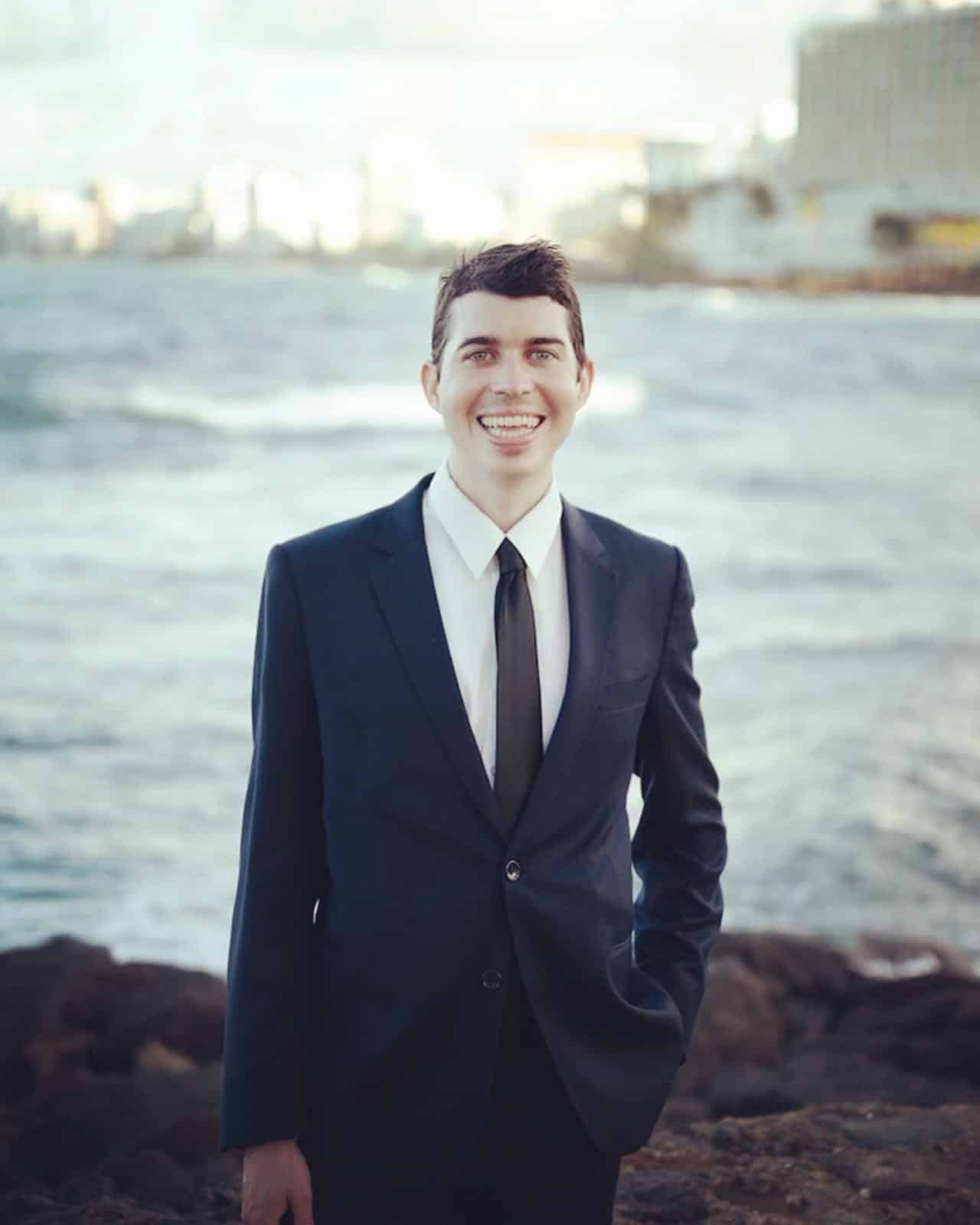 Alejandro smiles, in a dark suit, in front of a river.