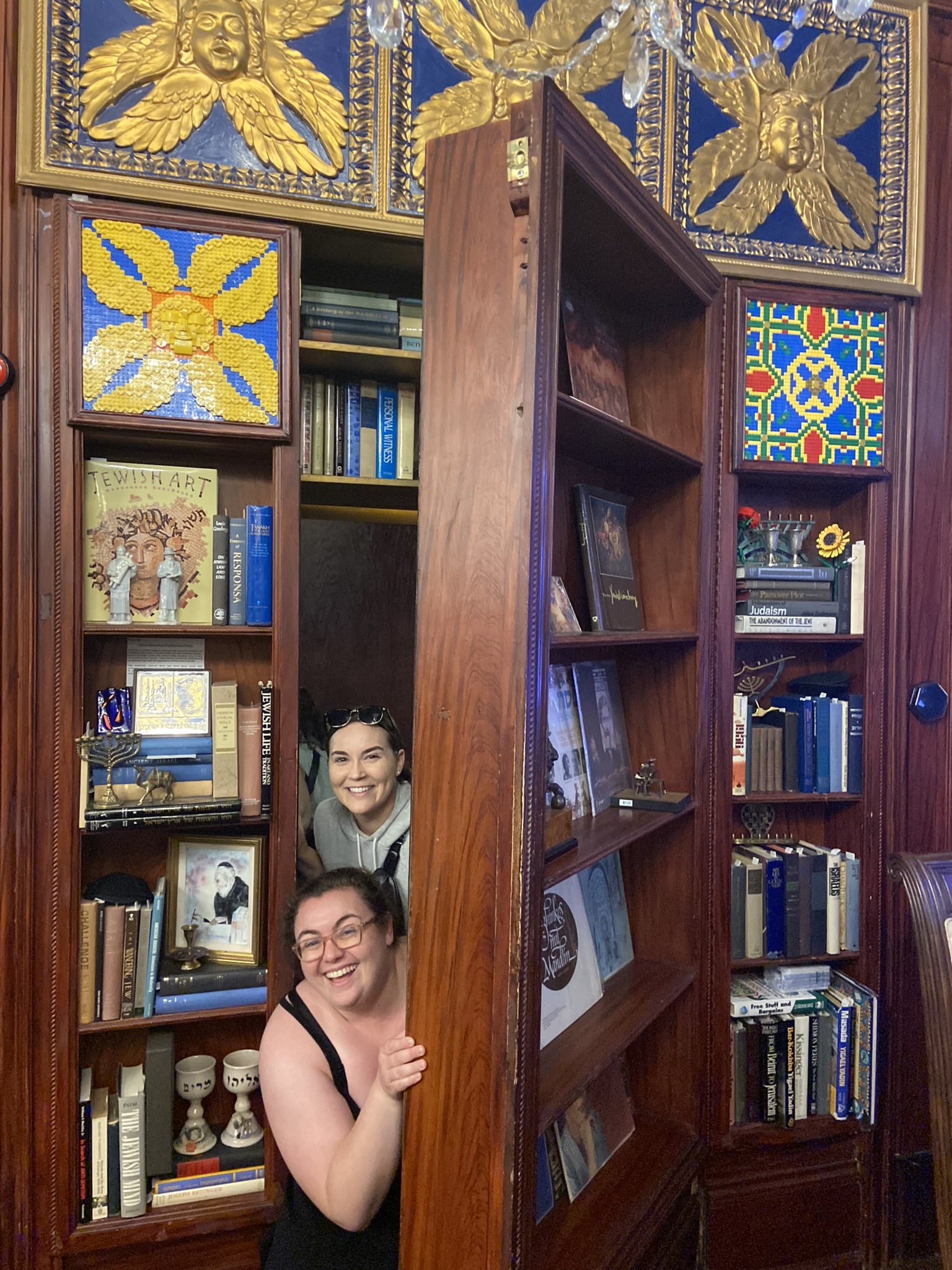 Miriam and a friend emerge from a hidden door in a bookcase.