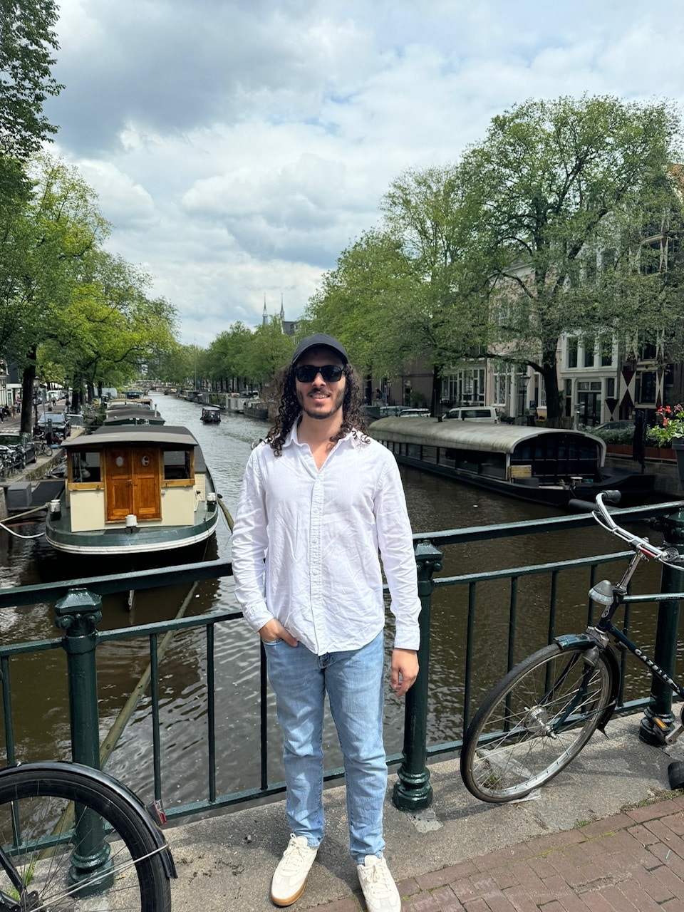 Eli stands on a bridge about a canal in sunglasses and a hat.