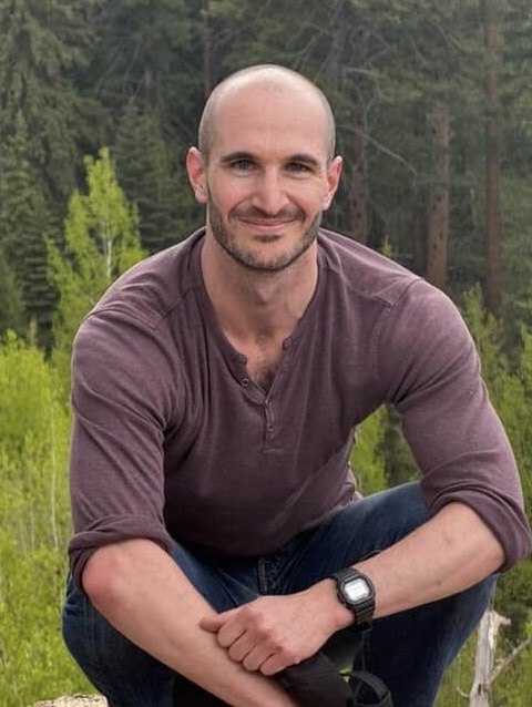 Aaron outdoors in a purple long sleeved shirt, kneeling.