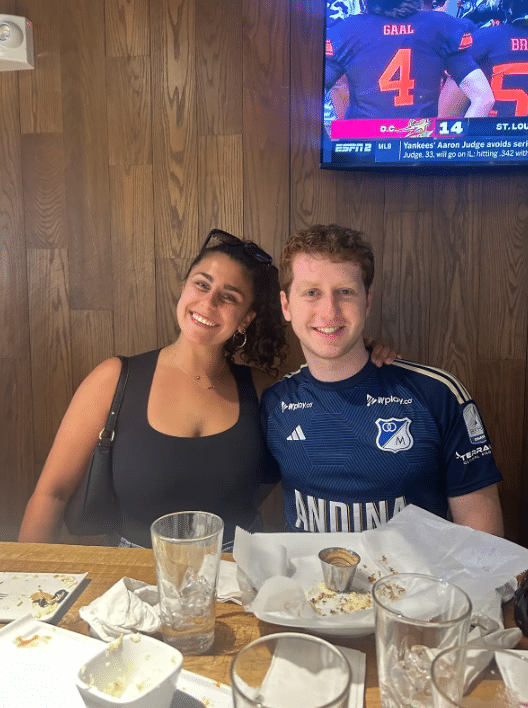 Maya having a meal with a man in a soccer jersey.